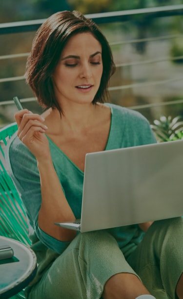 A woman holds an IQOS device while looking at a laptop.